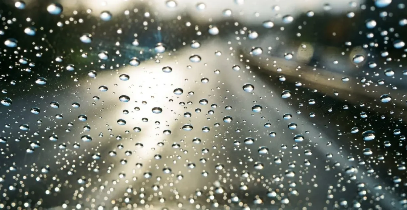 Nahaufnahme zeigt Lichtstreuung durch Mikrowassertröpfchen auf verschmierter Windschutzscheibe bei Gegenlicht