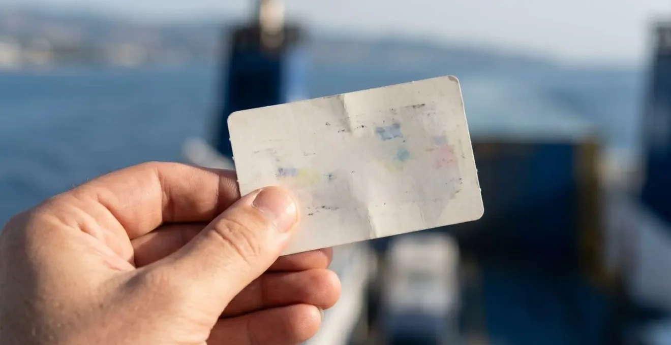 Nahaufnahme einer Hand, die ein Fährticket oder Smartphone mit verschwommenem Display hält, im Hintergrund unscharfes Meer und Schiff